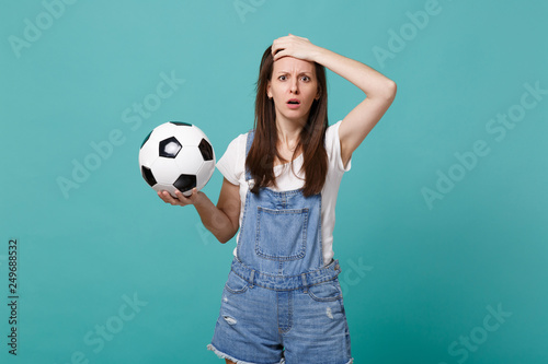 Shocked bewildered young woman football fan support favorite team with soccer ball putting hand on head isolated on blue turquoise background. People emotions, sport family leisure lifestyle concept. © ViDi Studio