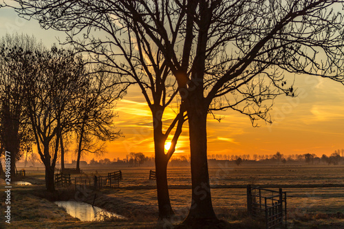 Sunrise over a rural landscape of frozen foggy grasslands, lines of canals, wooden fences and trees with farms on the horizon and an orange sky
