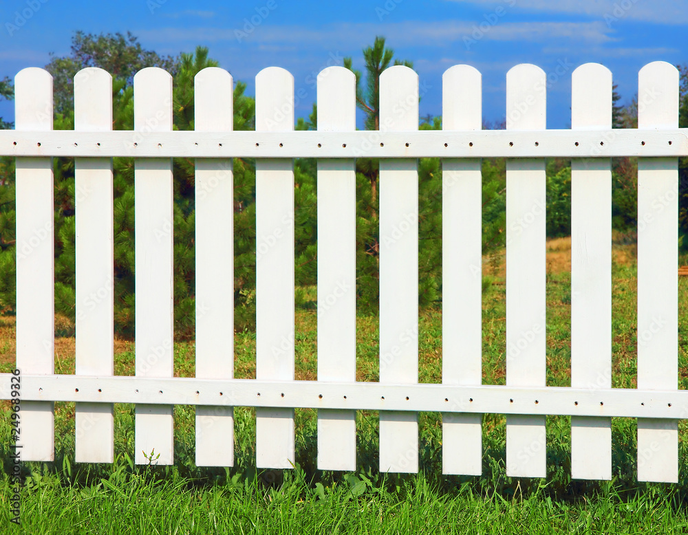 White wooden fence on green grass against of the trees and blue sky.