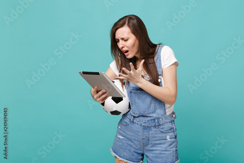 Preoccupied young woman football fan holding soccer ball, using tablet pc computer, spreading hands isolated on blue turquoise wall background. People emotions, sport family leisure lifestyle concept.