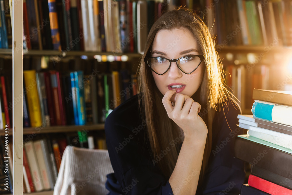 Beautiful young woman wearing glasses in the library among the bookshelves holding glasses and looking at the lens, reading, studying. Beautiful young woman wearing glasses in the library among the bookshelves holding glasses and looking at the lens, reading, studying.