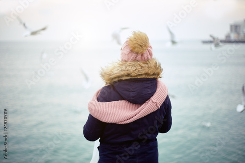 one girl in winter clothes are standing on the dock and feeding the gulls from their hands. Winter sea and birds.