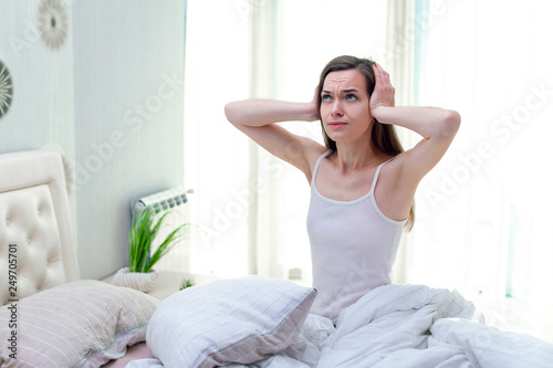 Young woman suffering and disturbed by noisy neighbors and covering her ears with hands while trying to sleep in bed at home in early morning