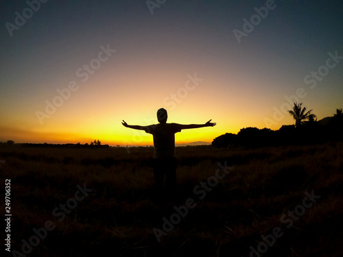 Silhouette Asian young man open arm is standing on grass city country home of field with sunset light and beautiful sky twilight sky and cloud. Success, happytime.