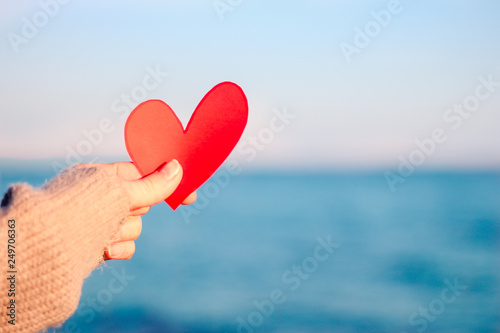 Valentine day concept, soft focus woman hand holding red heart, blurred sea background. Card with a carved red paper heart in a man's hand against a background of beautiful azure blue sea ocean.