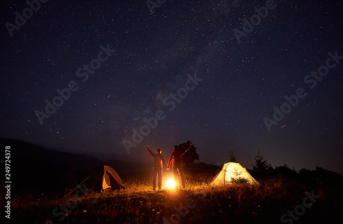Beautiful night camping picture. Bright bonfire burning between boy and girl standing with arms raised up in front of illuminated tents under amazing dark starry sky on distant hills background.