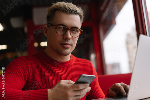 Handsome man in stylish eye glasses using mobile phone and downloading application for online shopping with discount sales, cash back. Portrait of successful developer or programmer working project. 