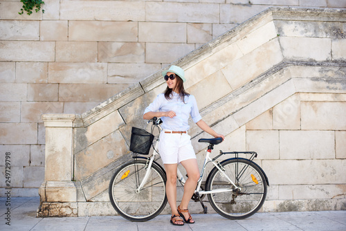Pretty smiling woman in sunglasses, white shorts, blouse and hat leaning on bicycle on white stone wall and stairs background on bright sunny day. Sightseeing and vacations concept.