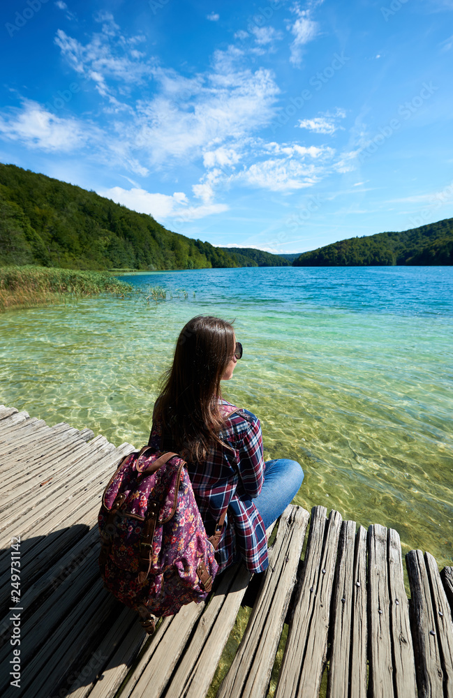 Back view of young tourist woman in sunglasses and with backpack sitting on wooden bridge enjoying beautiful view, colorful spring panorama of lit by sun green forest and blue lake under clear sky