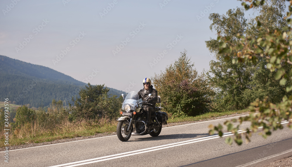 Bearded biker in helmet, sunglasses and black leather clothing riding cruiser powerful motorcycle down sunny asphalt road on bright summer day on background of green woody hill.