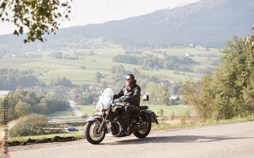 Long-haired bearded cool biker in sunglasses and black leather clothing riding cruiser powerful motorcycle along sunny asphalt road on bright summer day on background of rural misty landscape. © anatoliy_gleb