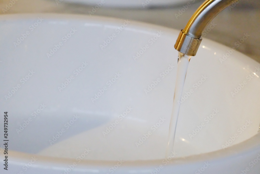 In selective focus water flowing from a steel faucet in a restroom with white ceramic sink background for saving water concept 