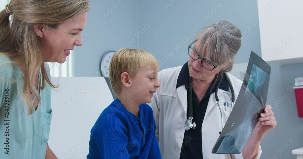 Senior woman pediatrician showing x-ray of spine to child patient and mom. Little boy at doctors office for scoliosis. Slow motion 4k Senior woman pediatrician showing x-ray of spine to child patient and mom. Little boy at doctors office for scoliosis. Slow motion 4k