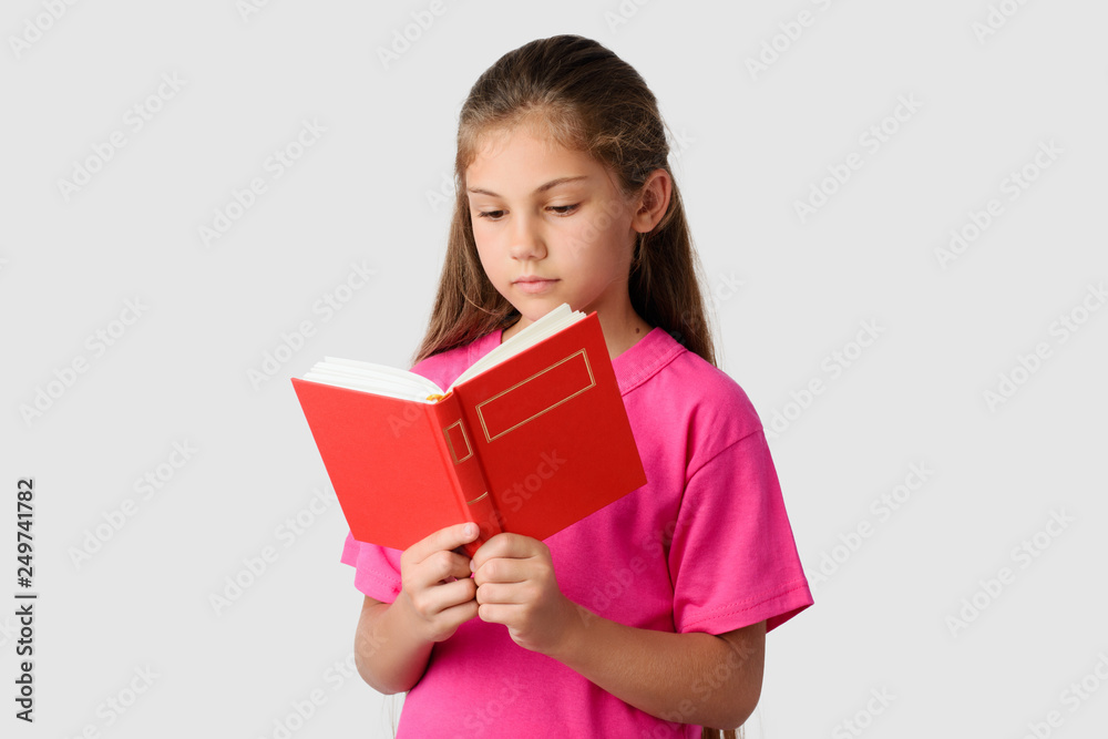 Smart little girl reading red book against a grey background. Lovely schoolgirl studying at home looking to the book attentively and learning material Smart little girl reading red book against a grey background. Lovely schoolgirl studying at home looking to the book attentively and learning material
