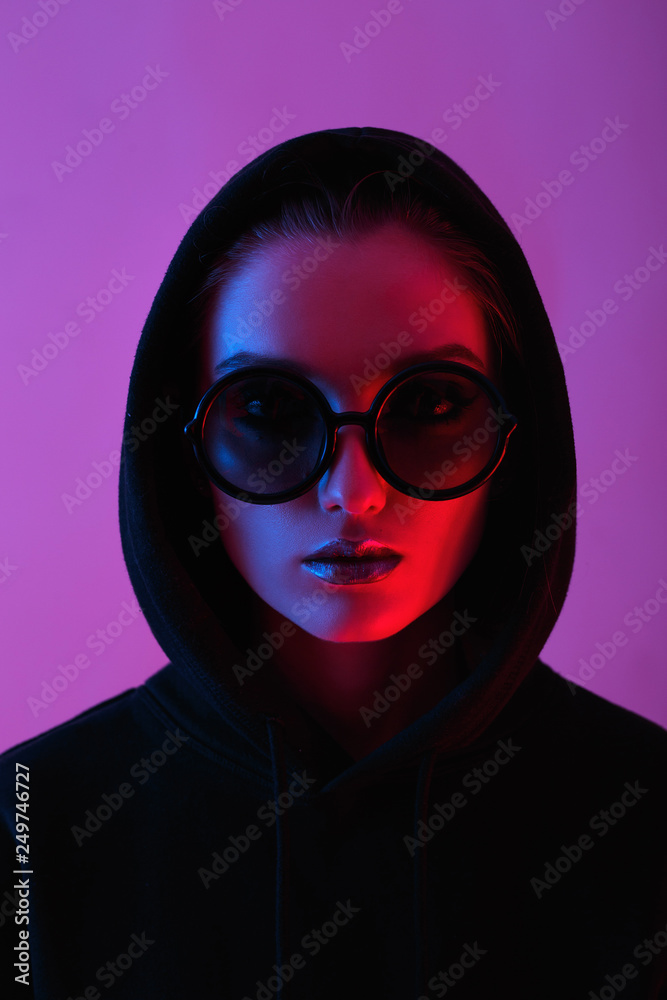 Portrait of fashion young girl in a black sweater with a hood and round sunglasses in red and blue neon light in the studio Portrait of fashion young girl in a black sweater with a hood and round sunglasses in red and blue neon light in the studio