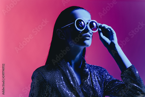 Fashion girl with long hair and round sunglasses in a black shining dress poses in neon light in the studio