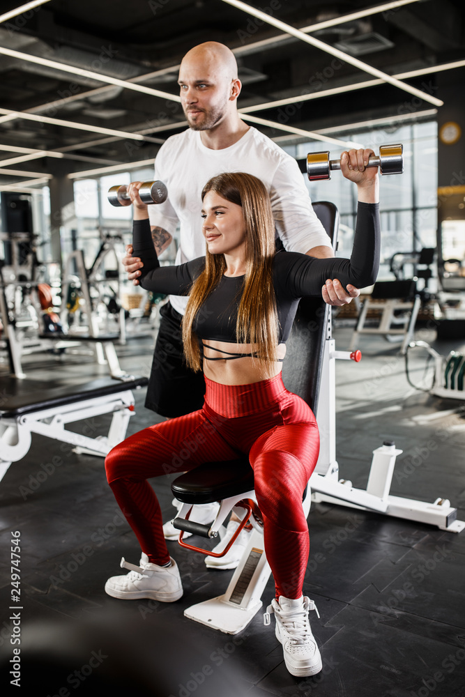 Beautiful slender girl in stylish sports clothes does a dumbbell bench press up sitting on a bench while the athletic man insures her in the modern gym Beautiful slender girl in stylish sports clothes does a dumbbell bench press up sitting on a bench while the athletic man insures her in the modern gym