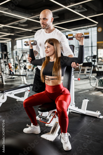 Beautiful slender girl in stylish sports clothes does a dumbbell bench press up sitting on a bench while the athletic man insures her in the modern gym