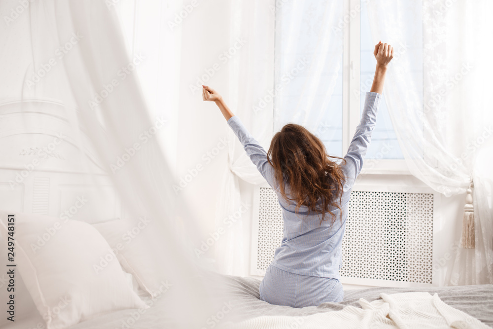 Brunette girl in the light-blue pajama stretches her arms up sitting on the canopy bed next to the window in the cozy bedroom - obrazy, fototapety, plakaty 