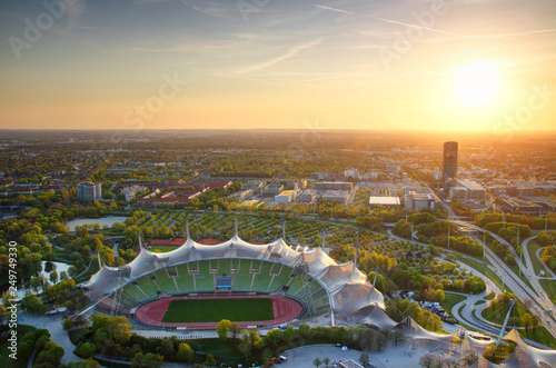Aerial view of modern European city outskirts lit by warm light of sun at sunset with commercial and sport facilities, parks, skyscrapers, tower blocks and highway junction, Munchen Bayern Germany