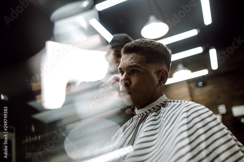The fashion barber in black clothes  makes a razor cut hair for a stylish black-haired man sitting in the armchair in a stylish barbershop