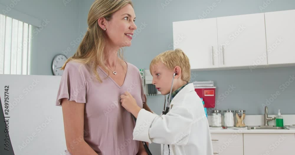 Cute little boy pretending to be a doctor with his mom while in pediatricians office using stethoscope. Child wearing oversized lab coat in exam room. Slow motion 4k Cute little boy pretending to be a doctor with his mom while in pediatricians office using stethoscope. Child wearing oversized lab coat in exam room. Slow motion 4k
