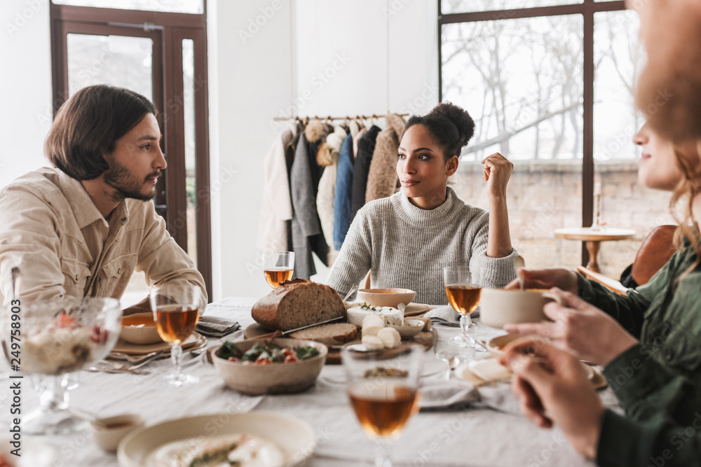 Pretty african american woman with dark curly hair sitting at the table thoughtfully looking at man with beard. Group of young international friends while spending time together on lunch in cafe