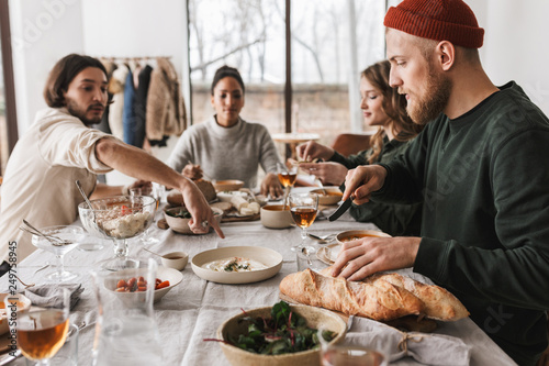 Handsome man in hat with beard sitting at the table with baguettes thoughtfully looking aside. Group of young international friends spending time together on lunch in cafe