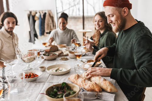 Handsome smiling man in hat with beard sitting at the table happily cutting baguettes. Group of young international friends spending time together on lunch in cozy cafe