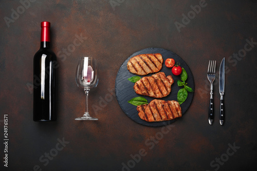 Billede på lærred Grilled pork steaks on stone with bottle of wine, wine glass, knife and fork on