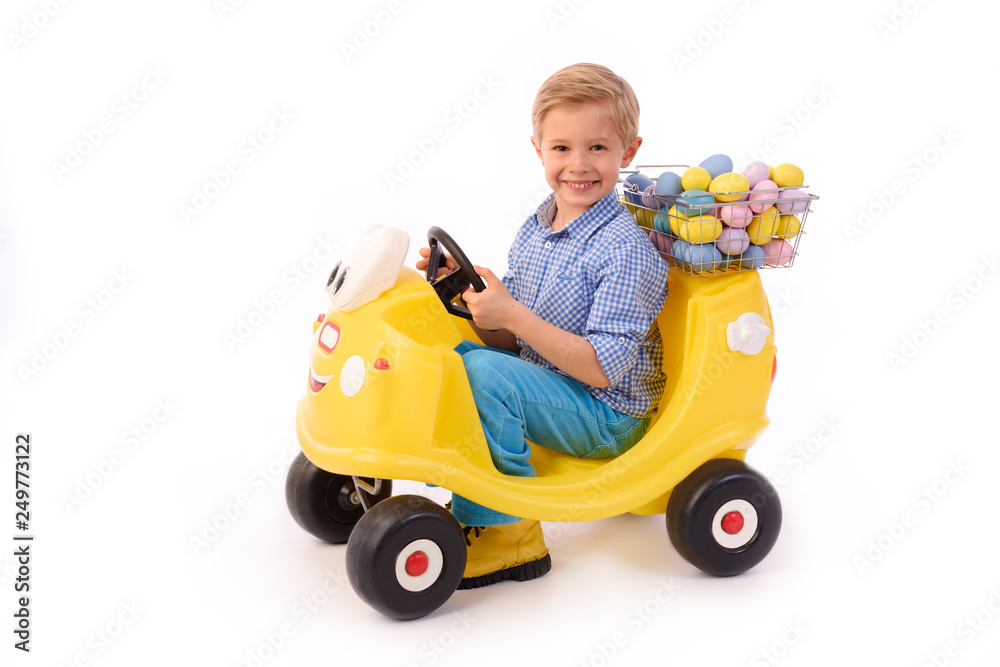Child and Easter. Cute little boy rides on a small car and carries colorful Easter eggs in a small basket. A boy dressed in blue, on a white background, studio. Facing camera.