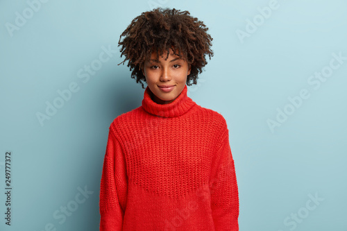 Headshot of beautiful young dark skinned woman with curly hair, dressed in red winter jumper, smiles pleasantly at camera, has no make up, expresses good feelings, isolated over blue background