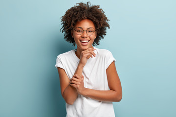 Isolated shot of happy joyful teenage girl with healthy dark skin, Afro haircut, holds hand under chin, hears positive calm story from friend, wears white shirt, isolated over blue background