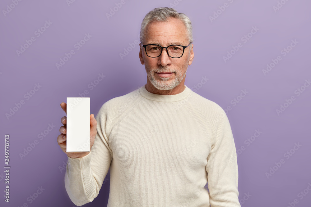 Isolated shot of handsome man with thick beard, grey hair, dressed in casual white jumper, holds vertical box with empty space, isolated over purple background. People and advertisement concept Isolated shot of handsome man with thick beard, grey hair, dressed in casual white jumper, holds vertical box with empty space, isolated over purple background. People and advertisement concept