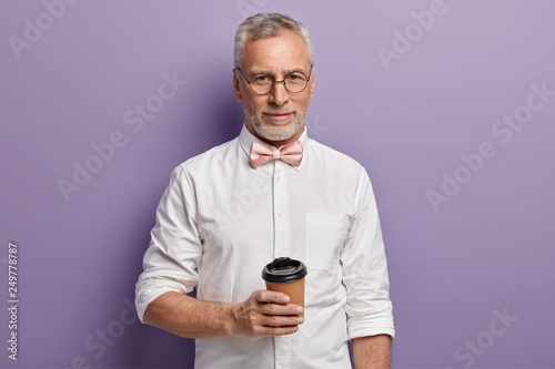 Shot of attractive elegant man with hot beverage, holds paper cup, wears elegant white shirt and pink bowtie, isolated over purple background, looks directly at camera. People, age, drink concept © Wayhome Studio