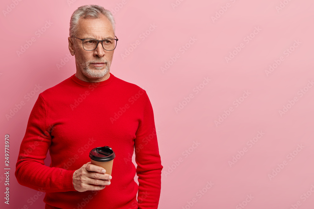 Indoor shot of pleasant looking serious elderly man in red clothing, holds takeaway coffee, focused aside, has break after work, isolated over pink background with blank space for your text. Indoor shot of pleasant looking serious elderly man in red clothing, holds takeaway coffee, focused aside, has break after work, isolated over pink background with blank space for your text.