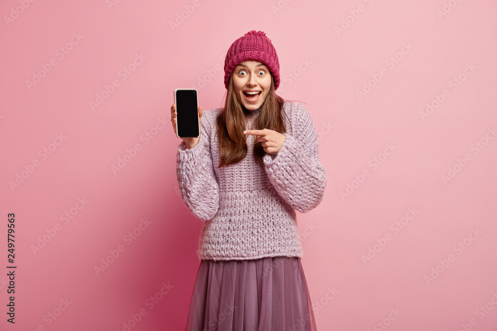 Buy this device. Carefree optimistic woman with happy expression, shows blank screen of mobile phone, wears loose sweater and headgear, models over rosy background, smiles broadly at camera. Buy this device. Carefree optimistic woman with happy expression, shows blank screen of mobile phone, wears loose sweater and headgear, models over rosy background, smiles broadly at camera.