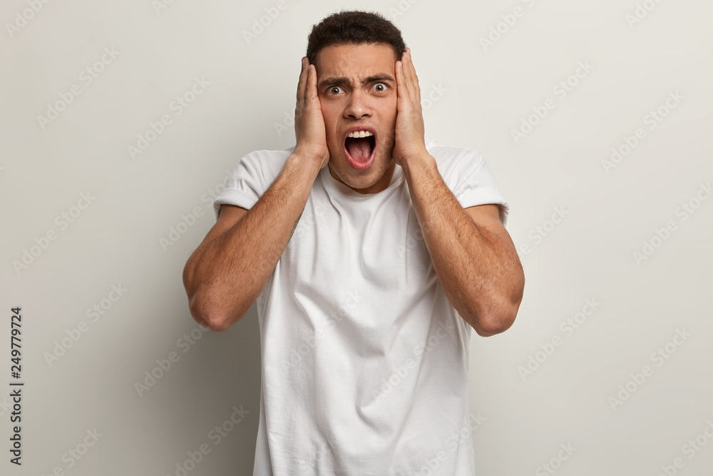 Depressed frustrated handsome guy keeps hands on head, opens mouth widely, exclaims from anger, being bothered, wears casual white t shirt, stands indoor. Annoyed youngster yells desperately Depressed frustrated handsome guy keeps hands on head, opens mouth widely, exclaims from anger, being bothered, wears casual white t shirt, stands indoor. Annoyed youngster yells desperately