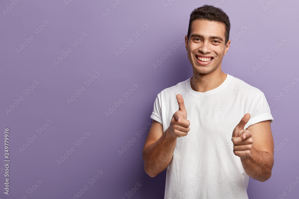 Photo of handsome smiling European man makes gun gesture, wears casual white t shirt, shows direction or expresses choice, models against purple background with empty space for your promotion