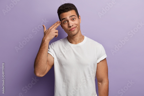Studio shot of attractive man with dark short hairstyle dressed in white t shirt points at temple, has muscular body, fed up with boring life, isolated over purple background. I am tired of everything