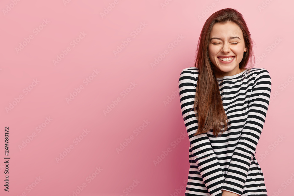 Portrait of pleased woman closes eyes from enjoyment, smiles broadly, shows white perfect teeth, wears striped casual black and white jumper, isolated over rosy background, free space for text Portrait of pleased woman closes eyes from enjoyment, smiles broadly, shows white perfect teeth, wears striped casual black and white jumper, isolated over rosy background, free space for text