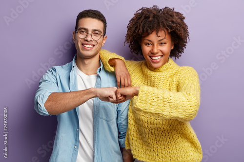 Mixed race partners give fist bump, agree to work together, have happy facial expressions rejoice success and cooperation, have toothy smiles, isolated over purple background. Team work concept
