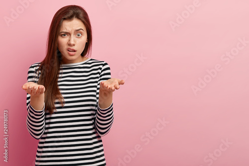 Indoor shot of young indignant woman gestures with indignation, frown face, wears striped sweater, cant understand why somebody treats her bad, isolated over pink background with blank space. © wayhome.studio 