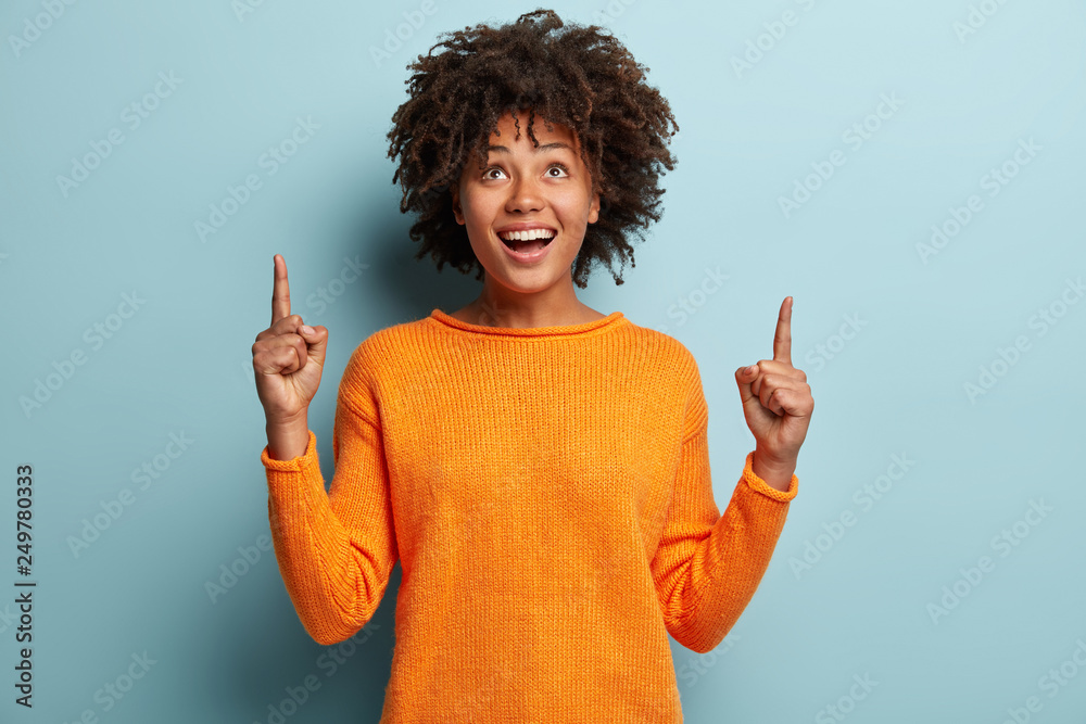 Horizontal shot of positive optimisitc African American woman points with both fore fingers upwards, wears casual jumper, advertises new item, isolated over blue background. Look at this thing - obrazy, fototapety, plakaty 