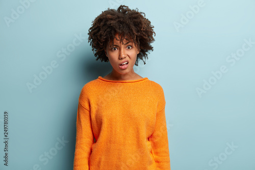Displeased woman with dark skin, Afro hairstyle, purses lips, feels discontet, looks with indignation, wears orange oversized jumper, isolated over blue background. Negative emotions concept