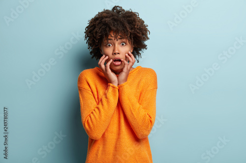 Photo of scared pretty woman keeps hands on cheeks, looks with surprisement and fear at camera, has curly Afro hairstyle, wears casual winter outfit, isolated over blue background. Omg concept