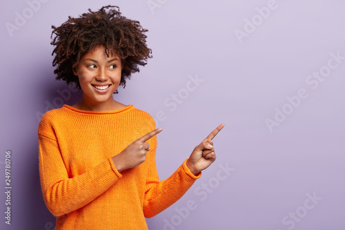 Shot of happy smiling black woman with Afro hairstyle, smiles positively, wears orange sweater, shows free space for your advertising content, isolated over purple background. Follow this direction