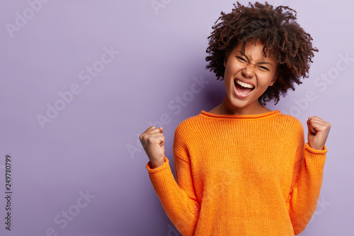 Life is wonderful! Joyful dark skinned lady keeps fists clenched, tilts head and exclaims in triumph, celebrates success, isolated over purple background with blank space for your advertisement