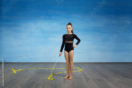 Girl gymnast in a black gymnastic swimsuit she prepared to do an exercise with a yellow gymnastic ribbon on a blue sky background.