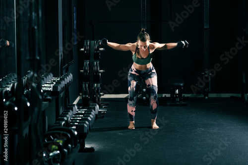 Strong muscular caucasian woman in sportswear and with pigtail lifting dumbbells barefoot. Gum interior.
